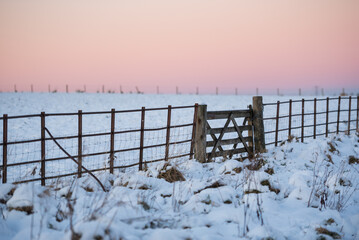 View of a fence with a gate under a red sky during sunset in a Winter day in the Pentland Hills Regional Park in Edinburgh, Scotland, UK, with the ground covered in snow