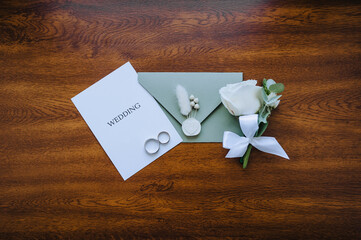 Wedding details and accessories close-up: rose boutonniere, gold rings, an envelope with a seal with a heart, a sheet of paper with an inscription. Photo, top view.