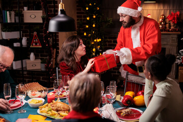 Man in santa claus costume greeting woman, giving christmas gift, wife getting present box from husband. Xmas night celebration, big diverse family eating traditional meal at festive dinner table