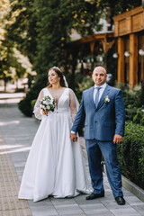 An adult stylish groom in a blue suit and a beautiful smiling bride in a blue dress are walking along the city street in the park, holding hands. Wedding photography of the newlyweds, portrait.