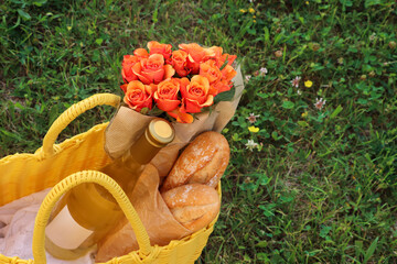 Yellow wicker bag with beautiful roses, bottle of wine and baguettes on green grass outdoors, above...