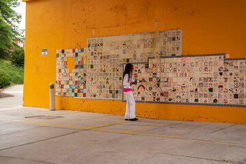 an African American woman with long sisterlocks wearing white and pink clothes, sunglasses and an orange head scarf  pointing to a wall mural on an orange wall holding a coffee cup with green trees - obrazy, fototapety, plakaty