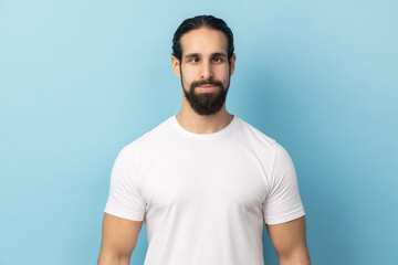 Obraz premium Portrait of comic positive man with beard wearing white T-shirt looking cross-eyed, having fun with silly face expression, playing fool. Indoor studio shot isolated on blue background.