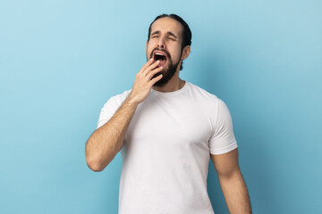 Portrait of drowsy tired man with beard in white T-shirt standing with wide open mouth, yawning feeling fatigued, lack of energy, need sleep and rest. Indoor studio shot isolated on blue background.