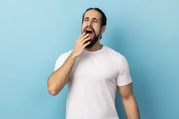 Fototapeta premium Portrait of drowsy tired man with beard in white T-shirt standing with wide open mouth, yawning feeling fatigued, lack of energy, need sleep and rest. Indoor studio shot isolated on blue background.