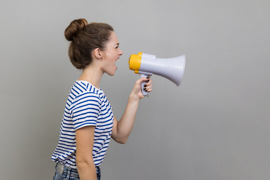 Side view of woman in striped T-shirt holding megaphone near mouth loudly speaking, screaming, making announcement, paying attention at social problems. Indoor studio shot isolated on gray background.