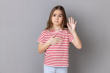 Portrait of serious patriotic little girl wearing striped T-shirt holding hand on heart, swearing...