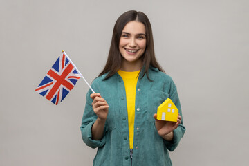 Portrait of smiling holding Great Britain flag and paper house, dreaming to buy accommodation in England, wearing casual style jacket. Indoor studio shot isolated on gray background.