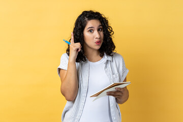 Portrait of amazed inspired woman journalist with dark wavy hair writing in paper notebook, having new excellent idea, raising finger up. Indoor studio shot isolated on yellow background. - obrazy, fototapety, plakaty