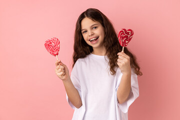 Portrait of satisfied little girl wearing white T-shirt holding two heart shape lollypops, looking...