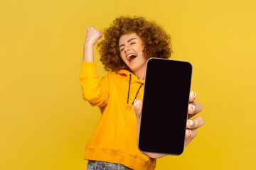 Amazed happy woman with Afro hairstyle wearing casual style hoodie showing big mobile display, clenched fist, celebrating victory, copy space. Indoor studio shot isolated on yellow background.