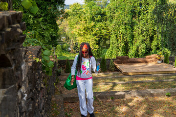 an African American woman with long sisterlocks wearing white and pink clothes, sunglasses and an orange head scarf carrying and green purse walking up a hill surrounded by lush green trees - obrazy, fototapety, plakaty