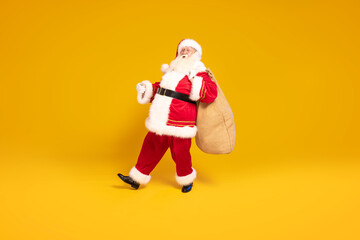 Fototapeta na wymiar Merry Christmas and a happy new year ! Real Santa Claus carrying heavy sack with gifts, standing on yellow studio background . Xmas time.