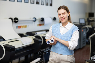 Portrait of positive woman printshop worker holding rolled pieces of paper and looking at camera.