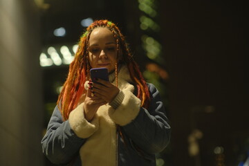 Young woman with dreadlocks browsing mobile phone at night in downtown.