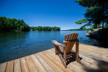 Adirondack chair on a wooden dock facing a lake in Muskoka. Cottages are visible across the water nestled between green trees.