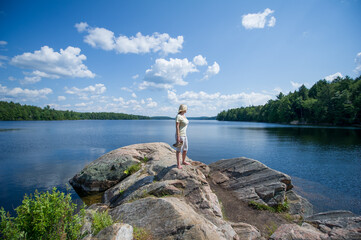 Young woman standing on a rock looking at Smoke lake in Algonquin Provincial Park, Muskoka Ontario Canada. The blue sky is showcasing beatiful fluffy clouds