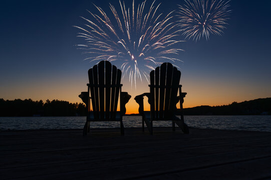 Canada Day fireworks over two Adirondack chairs  on the wooden dock in Muskoka, Ontario Canada, are facing the sunset orange hues  while facing the calm water.