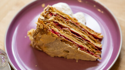 Trozo de torta de hojarasca, con interior de manjar y mermelada de frambuesa. Cubierto por una capa de crema merengue blanco. Sobre un plato de cerámica color morado