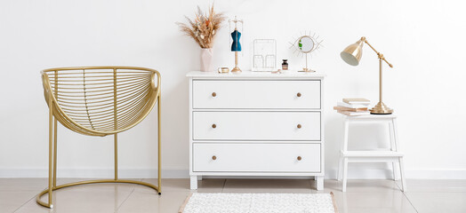 Chest of drawers with stylish jewelry and armchair near light wall in room