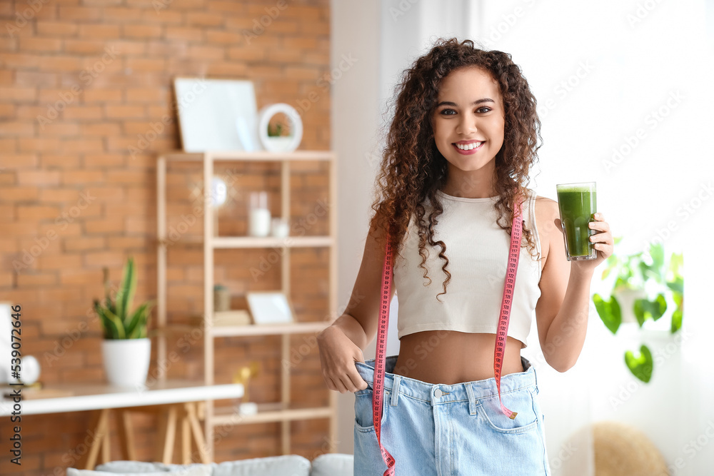 Wall mural happy young african-american woman in loose clothes, with smoothie and measuring tape at home. diet 