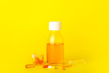 Bottle of cough syrup, cup, spoon and pills on yellow background