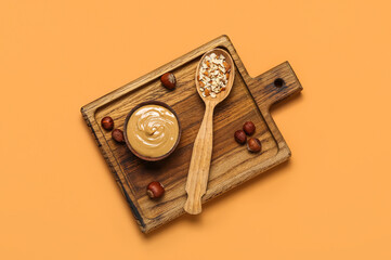 Wooden board with bowl of tasty nut butter and hazelnuts on color background