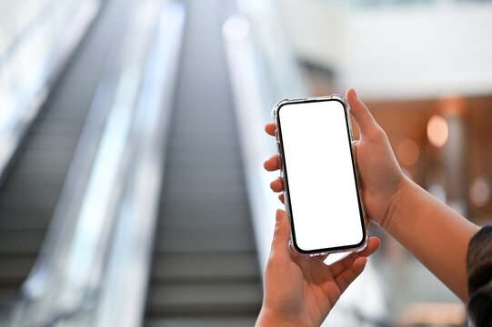 A smartphone mockup is in a woman's hands over blurred shopping mall's escalator