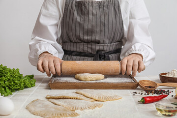 Woman rolling out dough for chebureks at table near light wall
