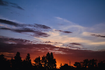 The morning glow illuminates the clouds on a beautiful colorful sky above the silhouettes of trees and a pillar at dawn