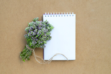 Empty paper notebook with purple thyme flowers bunch flat lay on wooden background. Bouquet of thyme flowers and blank notepad on background of craft paper