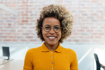 Portrait of smiling african american woman looking at camera with blonde curly hair in glasses in office. 