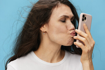 a funny, beautiful, pleasant woman with hair flying in the wind stands in a white T-shirt making a funny face and kisses her phone. Horizontal photo on a light blue background with empty space