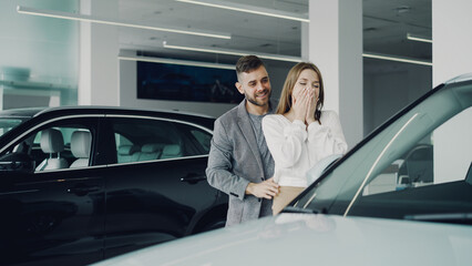 Fototapeta na wymiar Attractive young woman is getting new car from her loving boyfriend, he is closing her eyes and leading her to auto in motor dealership, they are hugging and kissing.
