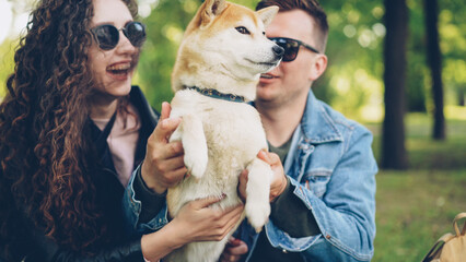 Joyful young people are playing with beautiful dog in the park having fun and laughing. Pretty girl with long curly hair is wearing leather jacket, guy is in modern denim jacket.