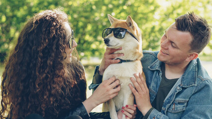 Cheerful young couple is having fun with dog putting sunglasses on its eyes and laughing while spending weekend in the park. Nature, modern lifestyle and animals concept.