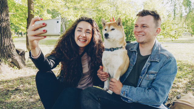 Proud dog owners happy couple are taking selfie with beautiful pet having fun and fussing it looking at smartphone. Modern lifestyle, animals and modern technology concept.