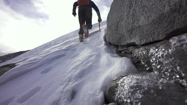 Person hiking up a snowy hill with crampons and ice axe, with partially frozen water in foreground