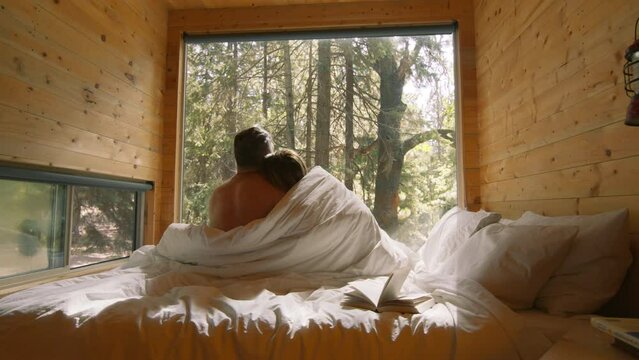 Authentic back view shot of young happy married couple of two tourists enjoying time together, hugging as sign of timeless love in a bed at small eco hotel with wooden cabins in spruce mountain forest