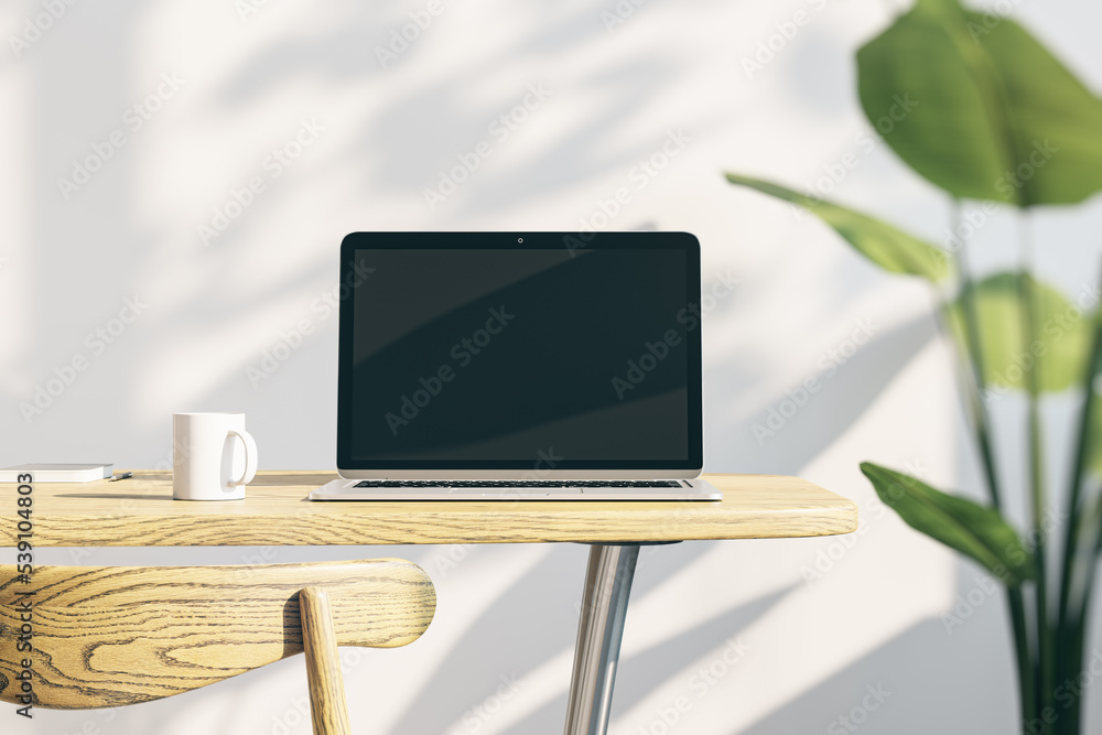 Poster designer office desktop with empty laptop screen with mock up place in frame, concrete wall background with sunlight and shadows, blurry decorative plant and coffee cup. 3D Rendering.
