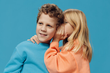 cute, beautiful children, brother and sister stand in bright clothes on a blue background and the girl tells secrets in the boy's ear. Studio photography with blank space for advertising mockup