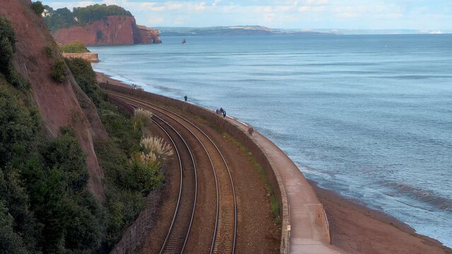 GWR Class 800 Penzance to Paddington train passes slowly along the stunning South Devon sea front between Teignmouth and Dawlish as people walk on the South West Coastal Path