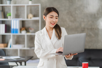 Cheerful young asian businesswoman holding digital tablet smiling and looking at camera standing in modern office. Wireless technology concept