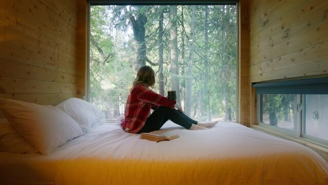 Woman on outdoor adventure trip in mountain rustic cabin with amazing forest view. Traveler in plaid shirt sitting on bed with cup of tea, looking at the window and reading a book, dreaming about life