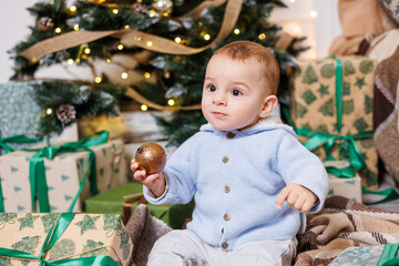 A one-year-old boy sits near a decorated Christmas tree with gifts. Christmas tree in the house. A happy child is waiting for the new year