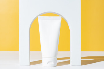 Blank white plastic cosmetics tube and white arch podium on yellow background. Skincare, beauty treatment, spa concept. Trendy showcase of cosmetics products