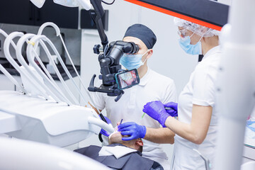 A dentist treats teeth using a dental microscope and tools. The assistant holds a syringe with air and water. Dental office