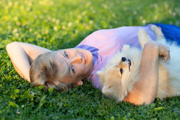 adult woman lying on grass and hugging dog. happy middle age lady with white Pomeranian German spitz. female owner with pet. concept of friendship, love, devotion