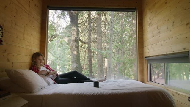 Beautiful Relaxed Woman Reading A Book While Laying On Bed At Large Window With Great View On Green Mountain Forest On Sunny Summer Day. Weekend Trip Or Escape To Wild Nature Concept. Nature Travel 4K