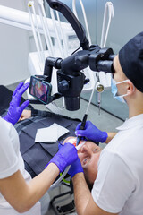 A young dentist looks at the patient's teeth with a dental microscope and holds dental instruments near the mouth. An assistant helps the doctor. They wear white uniforms with masks
